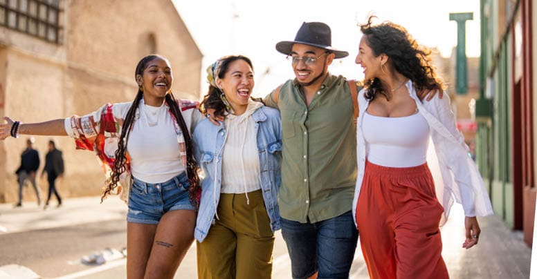 A cheerful diverse group of four friends walk down a city street, laughing and engaged in conversation, dressed in casual attire suitable for a relaxed day out.